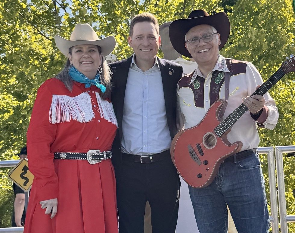 Three people smiling, wearing cowboy hats, one holding a guitar, outdoors with trees.