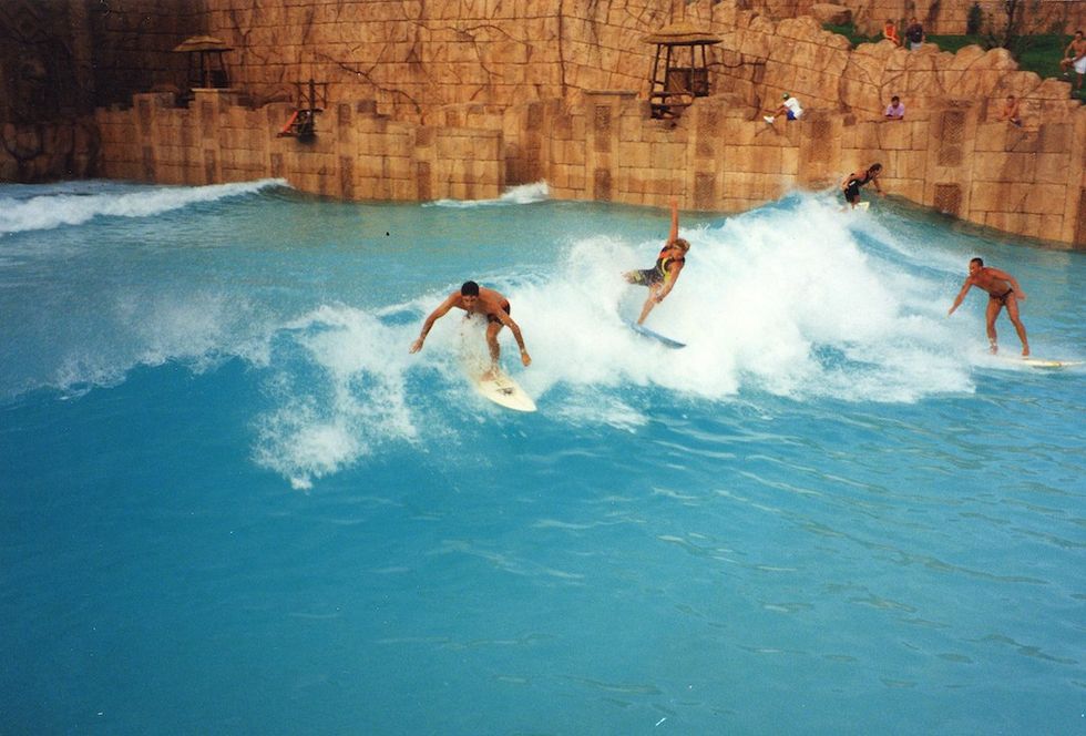 Three people surfing on artificial waves in a pool near rocky walls.