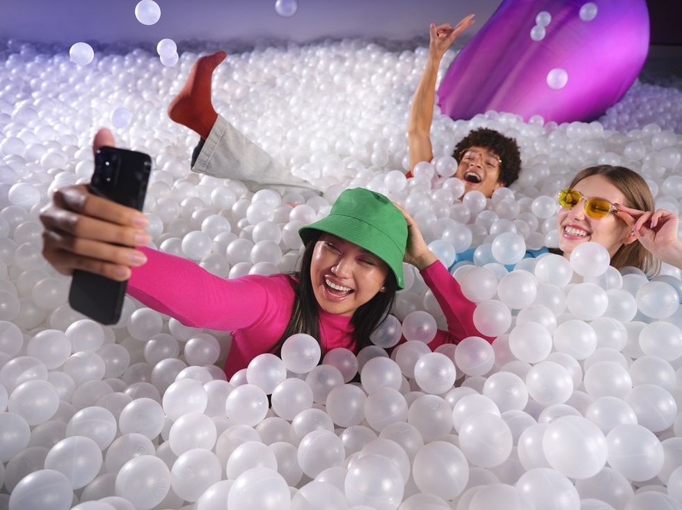 Three people take a selfie while submerged in a colorful ball pit, smiling and playful.