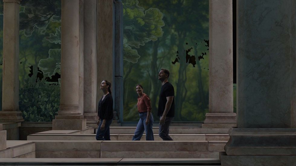 Three people walk through a columned area with large, leafy wall art.