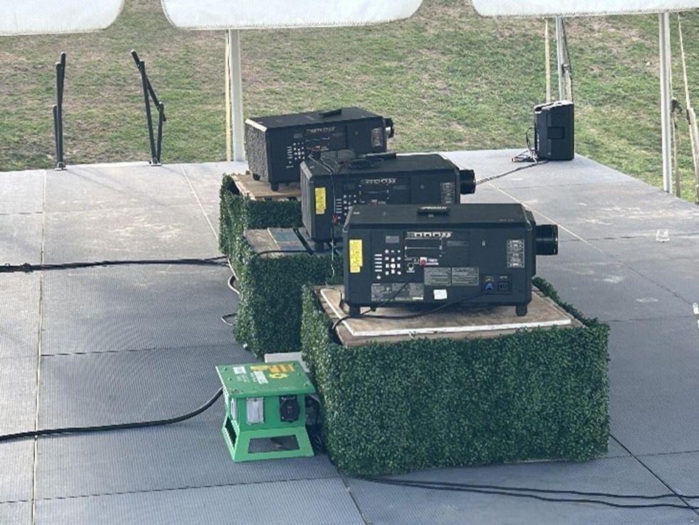 Three projectors on grass-covered stands under a canopy on a stage.