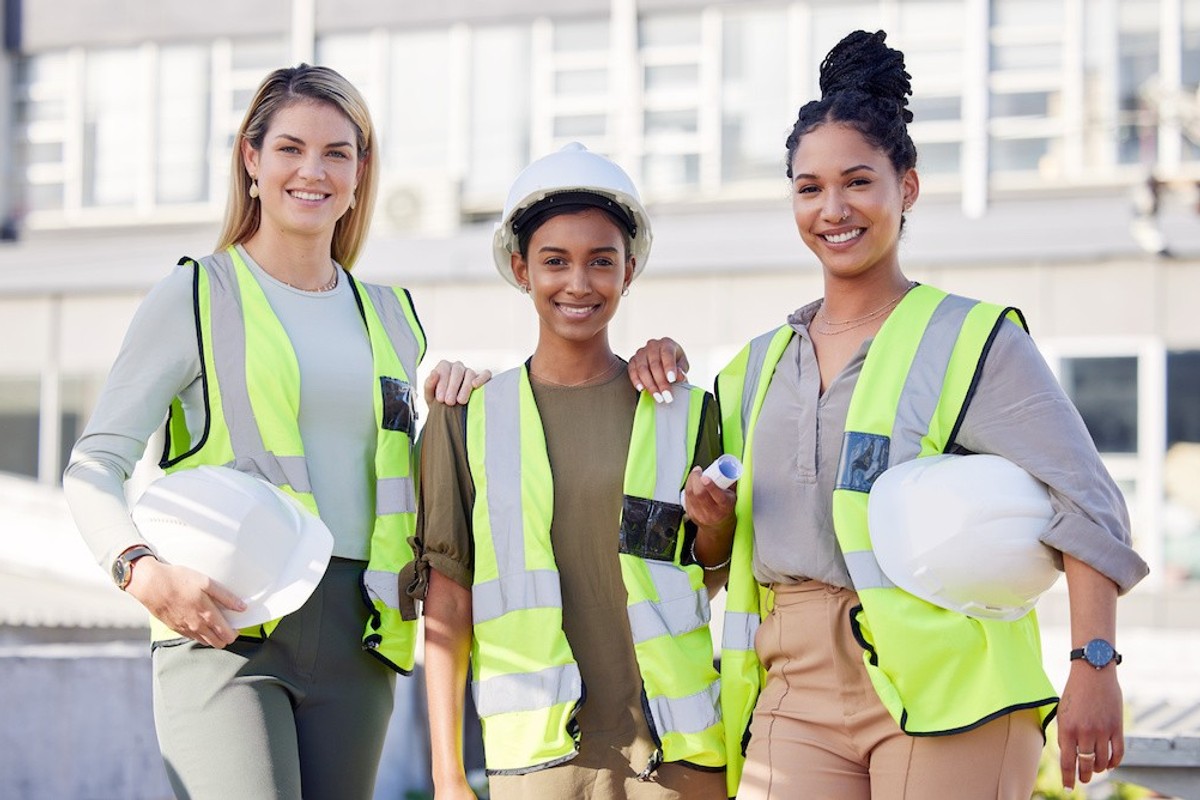 Three women in safety vests and helmets smiling at a construction site.