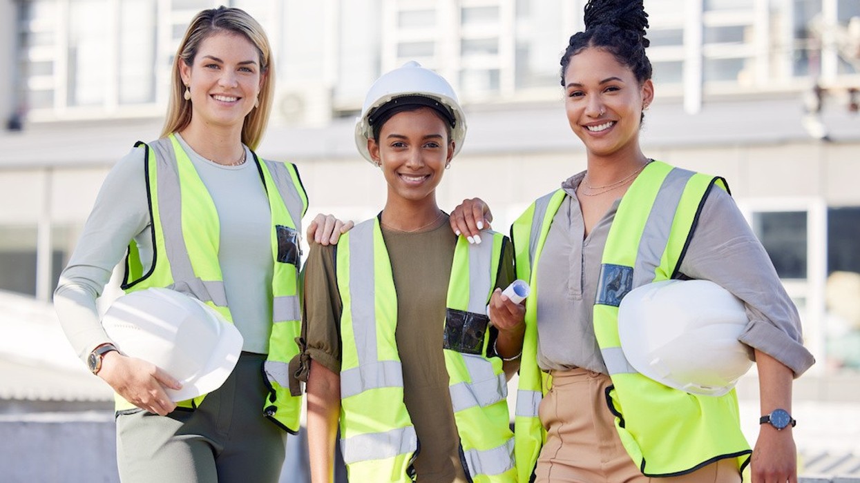 Three women in safety vests and helmets smiling at a construction site.