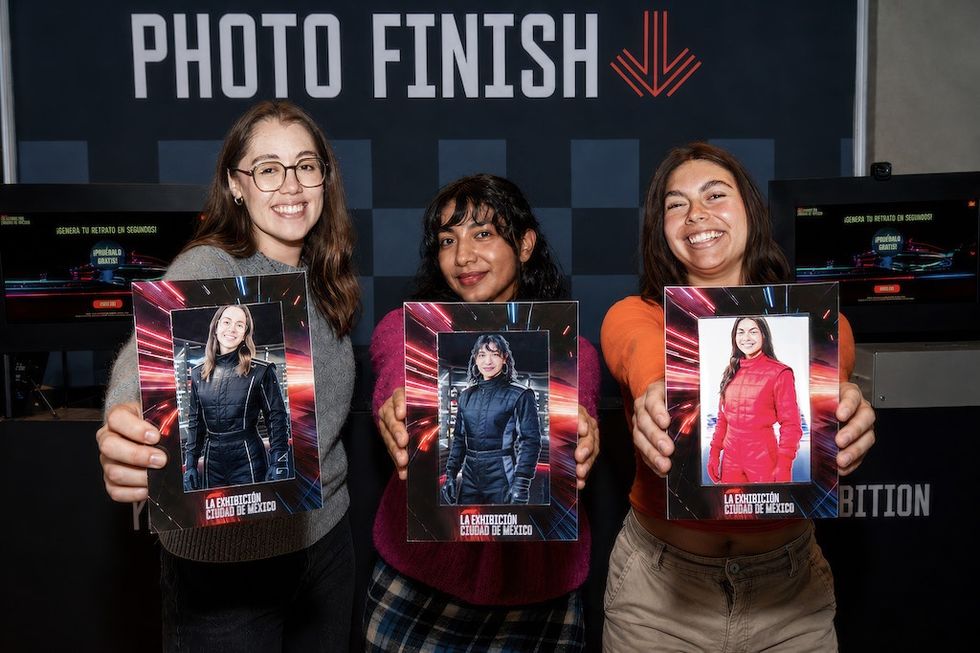 Three women smiling and holding racing-themed souvenir photos at Formula 1 La Exhibici\u00f3n