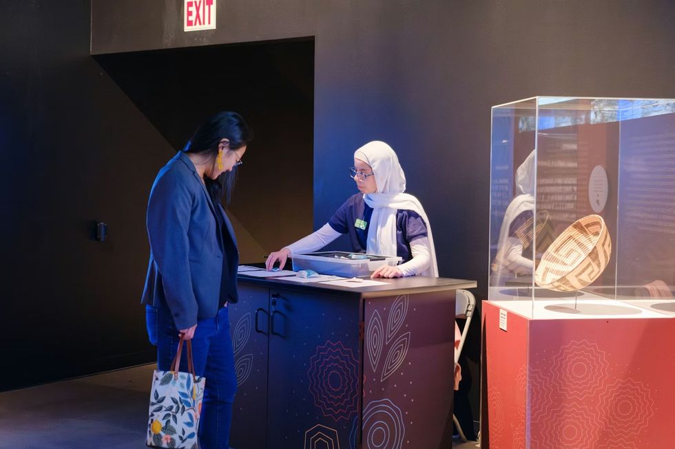 THRIVE \u2014 The First People: Yesterday, Today, and Forever. Two women interacting at a museum information desk, next to a woven basket display.