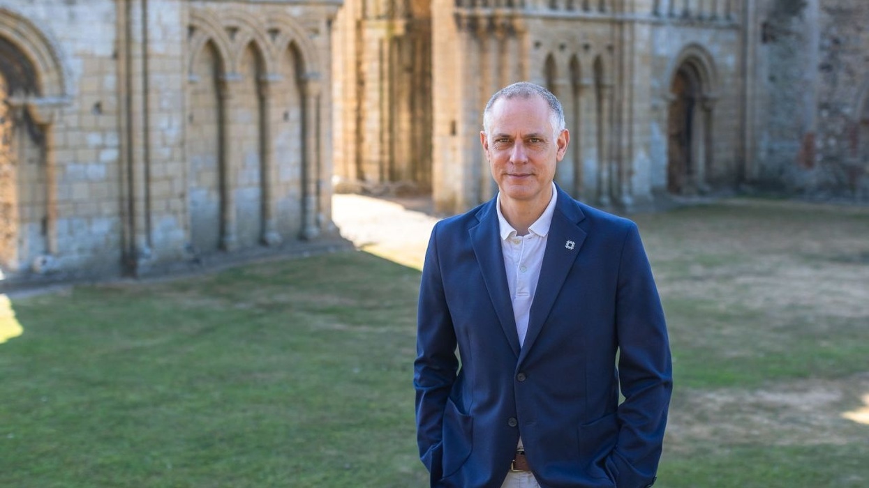 Tim Reeve in blue blazer standing near historic stone building, grassy area in foreground.
