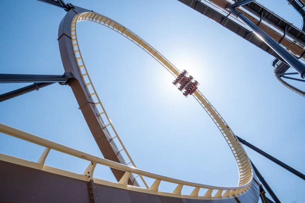 Tormenta: Rampaging Run roller coaster in a loop against a clear blue sky, with sun shining behind the riders.