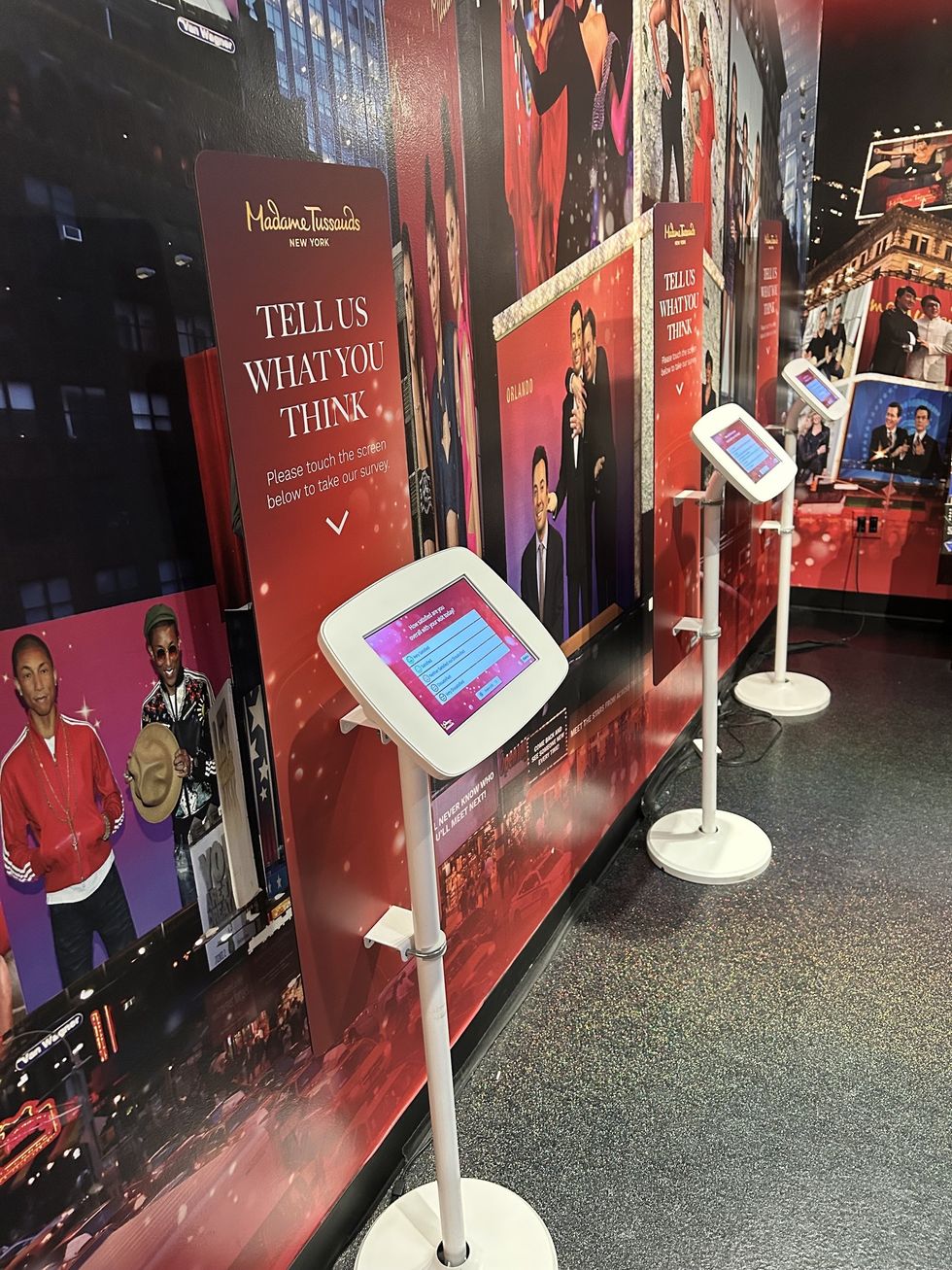 Touchscreen survey kiosks at Madame Tussauds New York, with celebrity wax figures in the background.