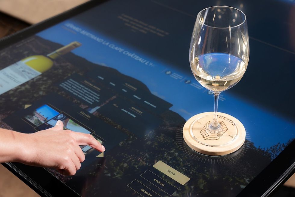 Touchscreen with wine info, glass of white wine on coaster, hand interacting.