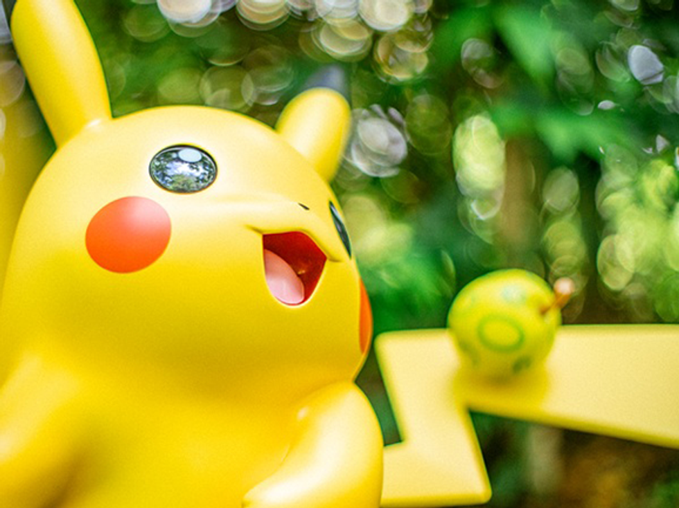 Toy Pikachu smiling with a small green fruit on its tail, set against a blurred leafy background.