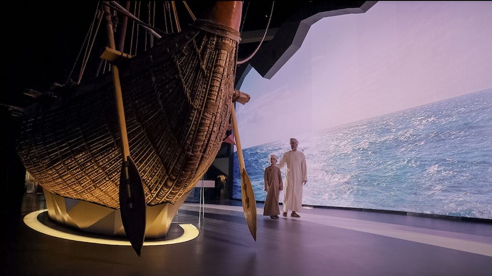 Traditional boat display with two people walking, ocean background on screen.