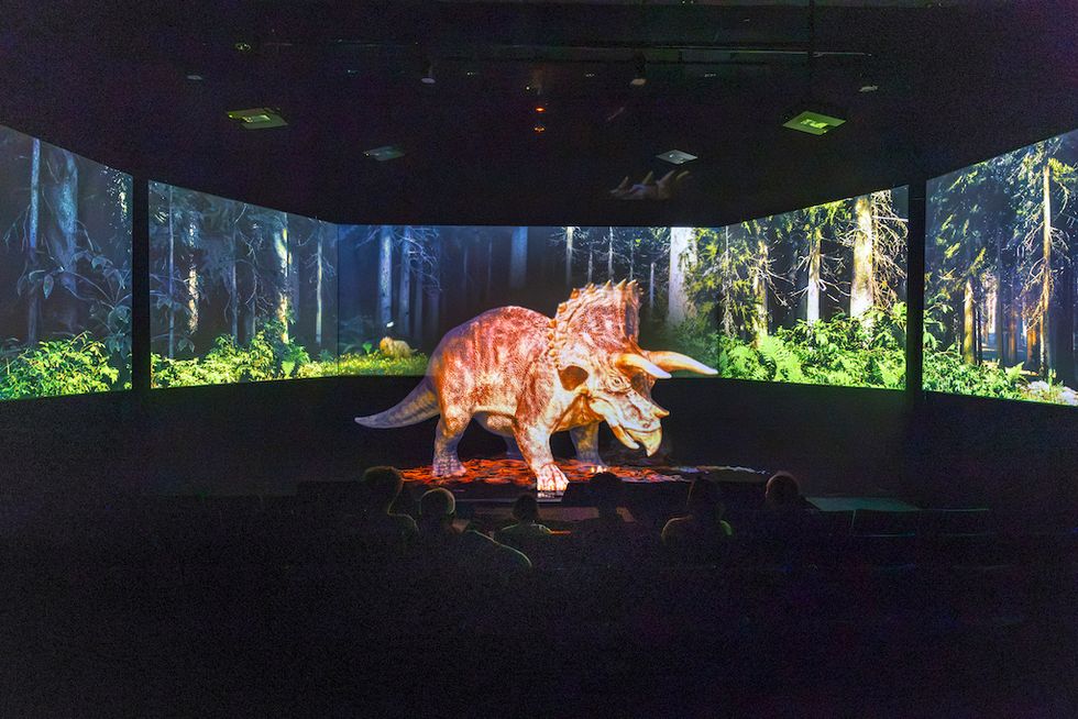 Triceratops hologram in a forest-themed exhibit room with seated viewers.