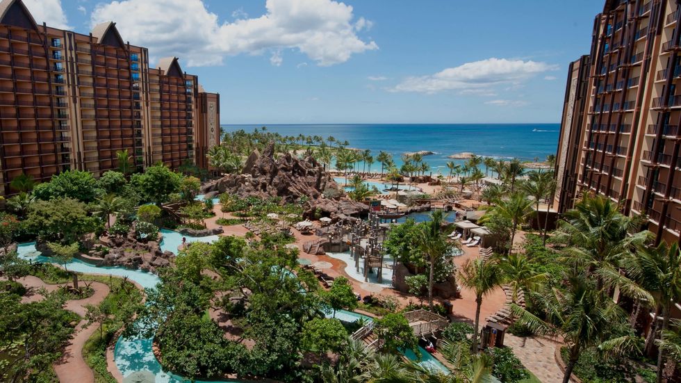 Tropical resort called Aulani, A Disney Resort & Spa with pools, gardens, palm trees, and ocean view under a clear blue sky.