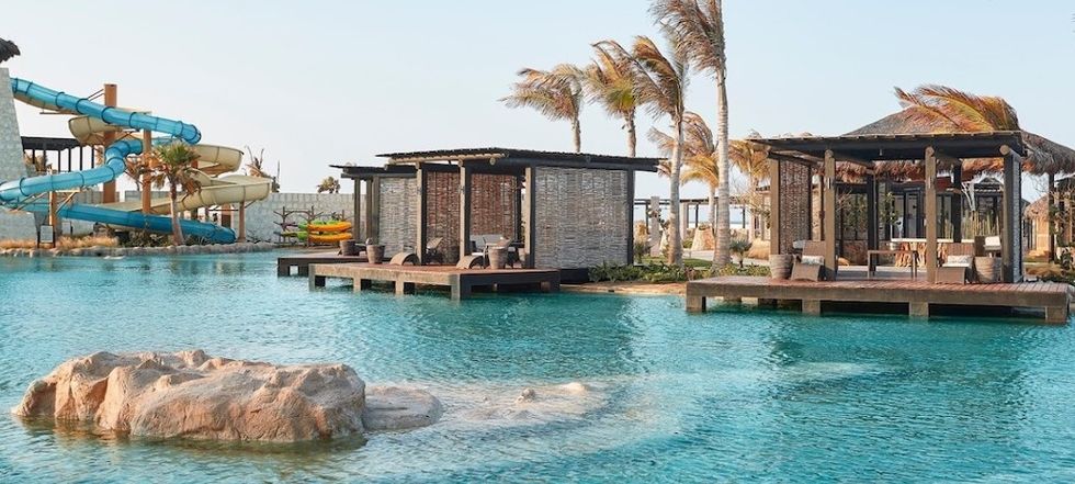 Tropical resort pool with cabanas, waterslide, and palm trees.