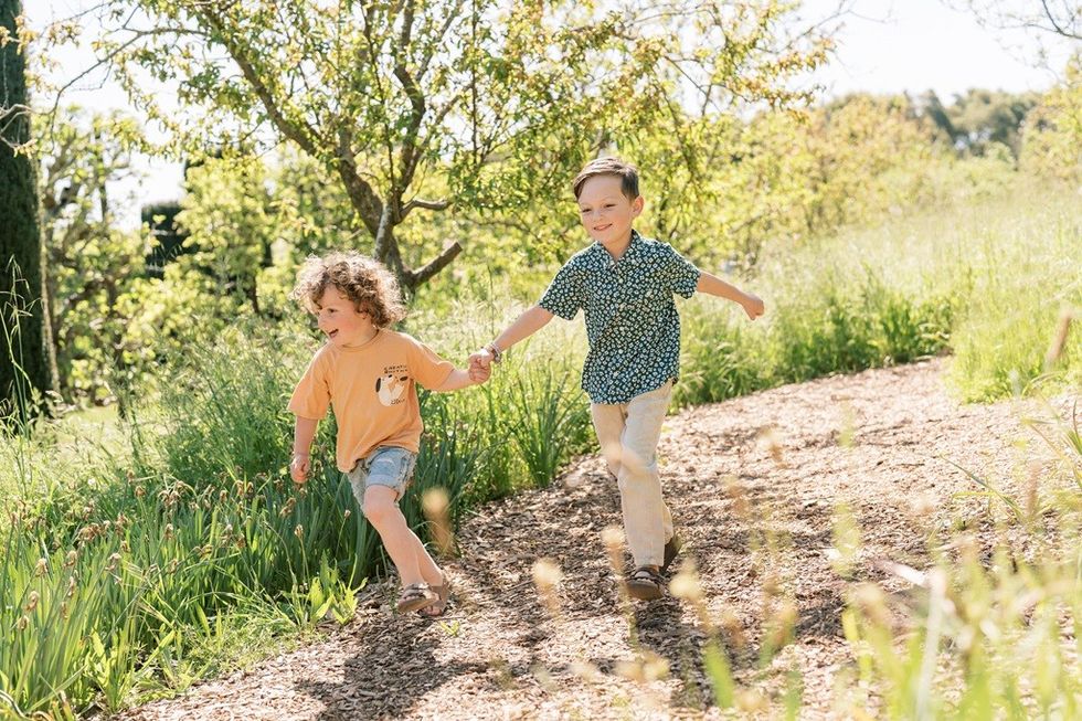 Two children joyfully run hand in hand on a garden path amidst greenery.