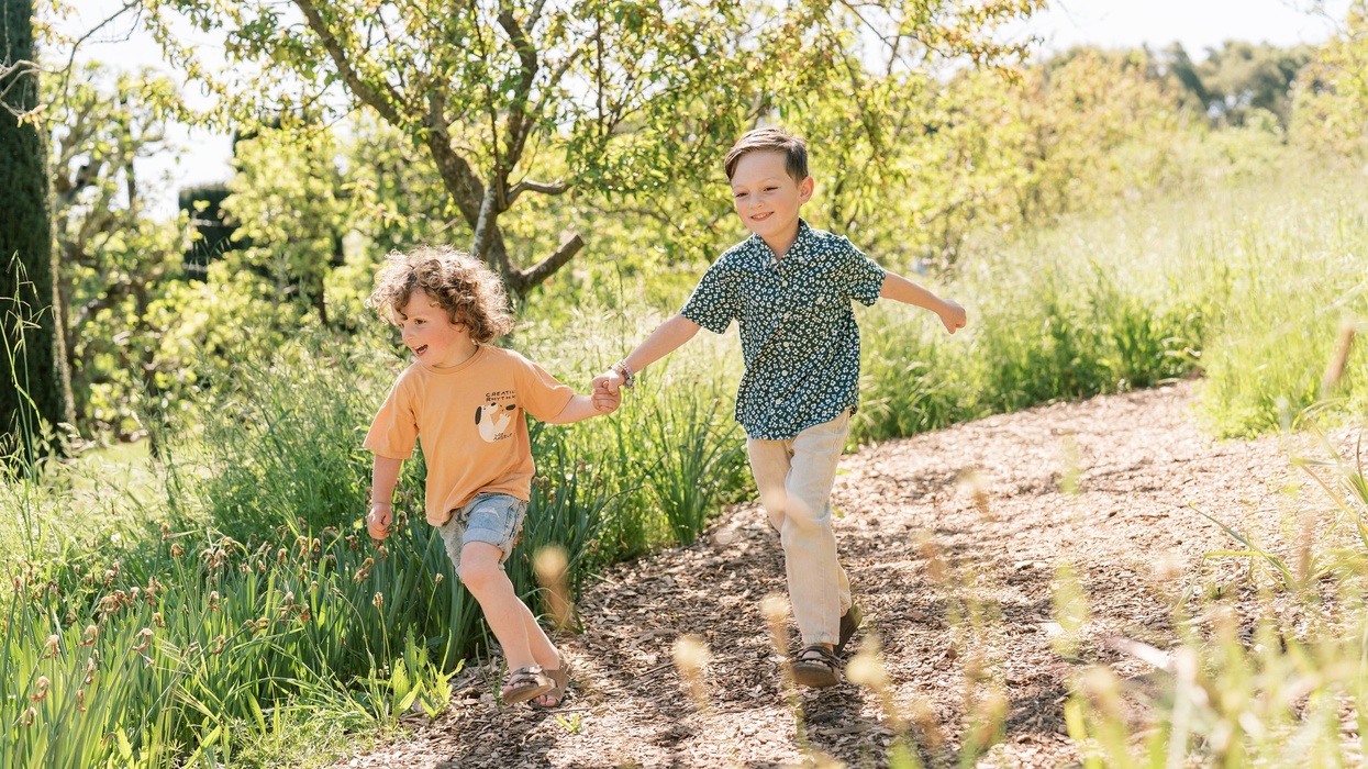 Two children joyfully run hand-in-hand on a sunlit, tree-lined path.