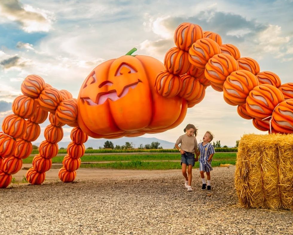 Two children walk beneath a giant jack-o\u2019-lantern arch made of pumpkins at a farm.