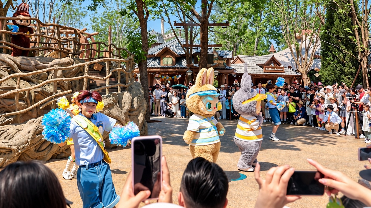 Two costumed rabbit characters and a cheerleader perform for a crowd at a sunny theme park.