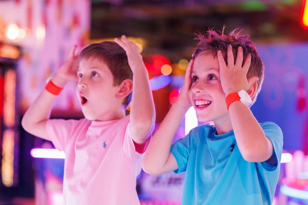 Two excited kids react with hands on heads in a colorful, neon-lit setting with PlayMAXX RFID bands