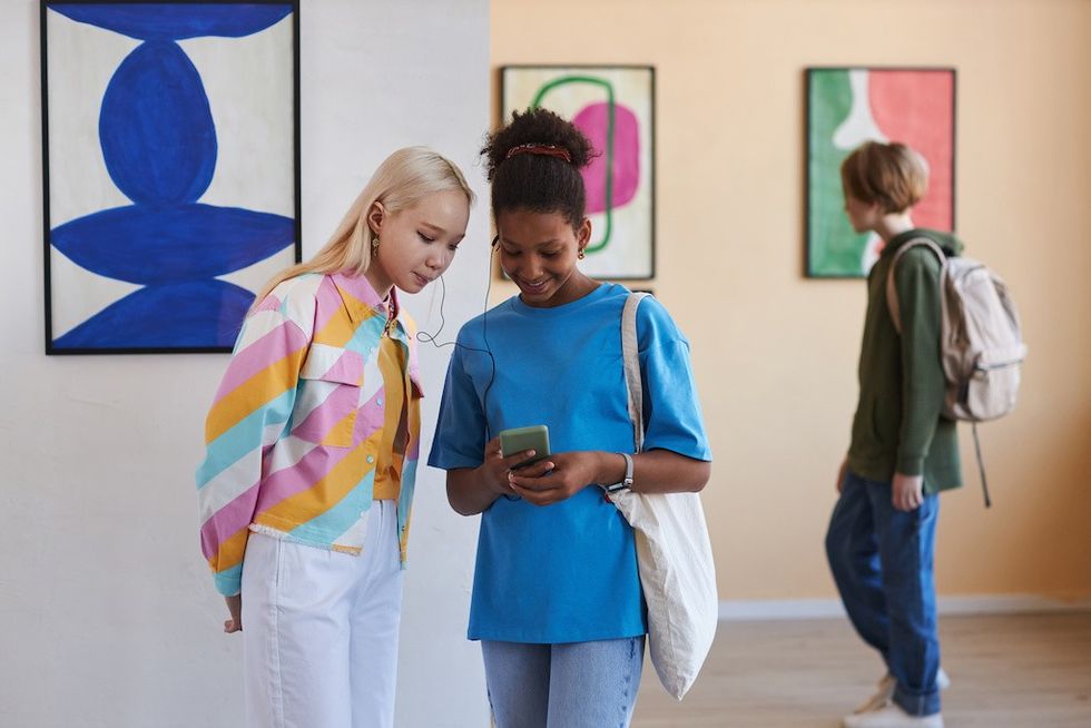 Two friends share earphones, looking at a phone, with paintings in the background.