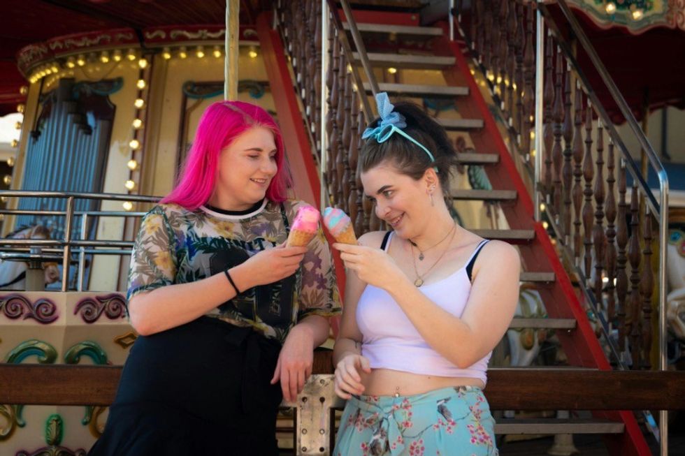 Two friends share ice cream cones in front of a carousel.