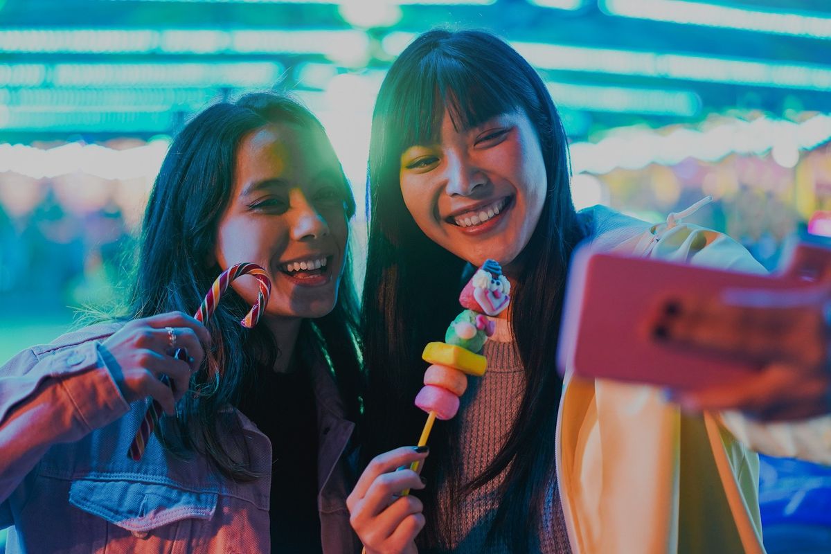 Two friends taking a selfie with candy at a bright, colorful carnival.