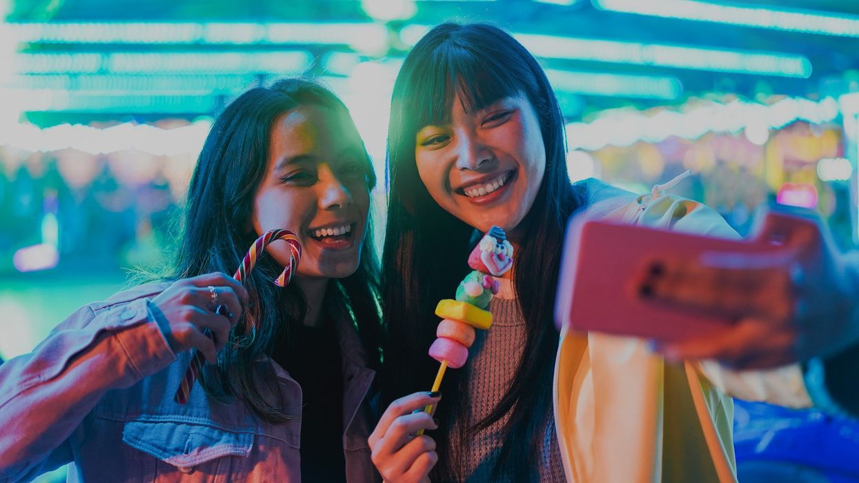 Two friends taking a selfie with candy at a bright, colorful carnival.