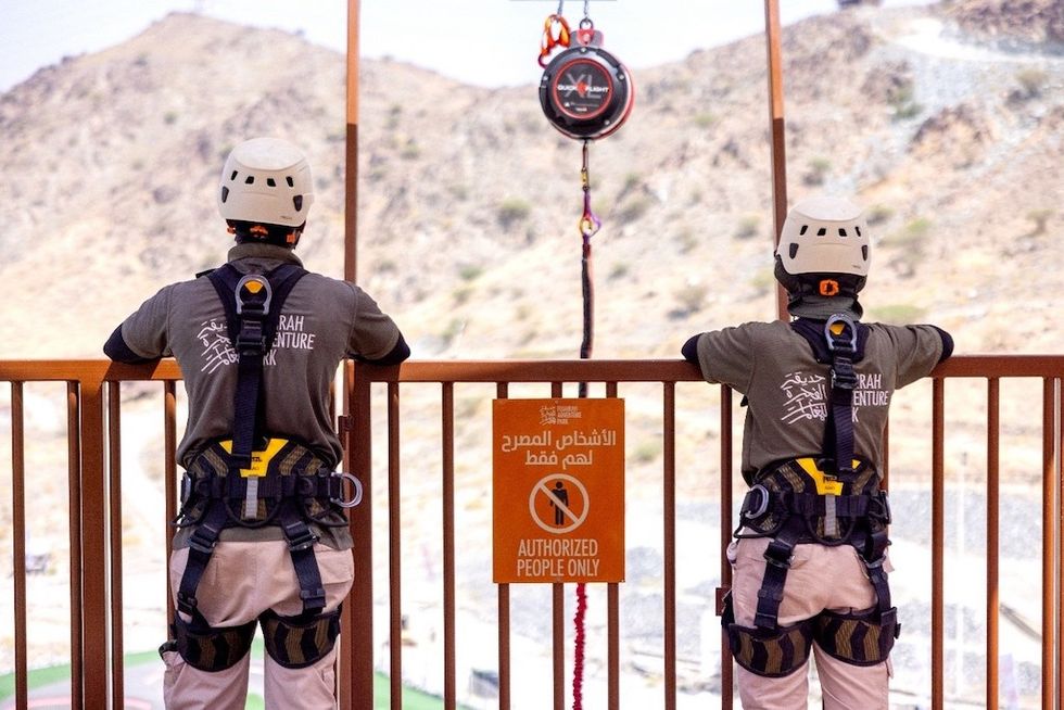 Two harnessed individuals stand by a railing, facing a mountain backdrop at Fujairah Adventure Park