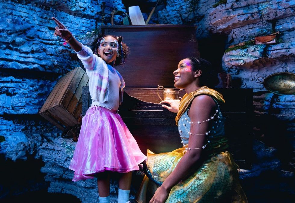 Two joyful kids in bright costumes pointing inside a cave-like setting.