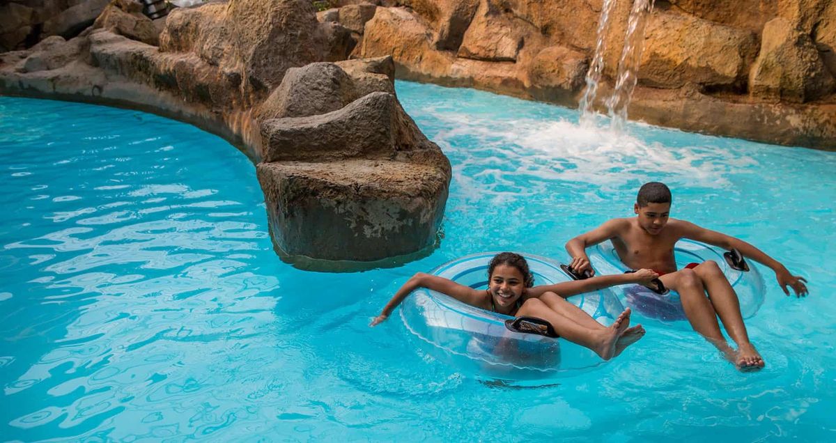 Two kids enjoying a lazy river ride in inflatable tubes near a rock waterfall at Alton Towers water park