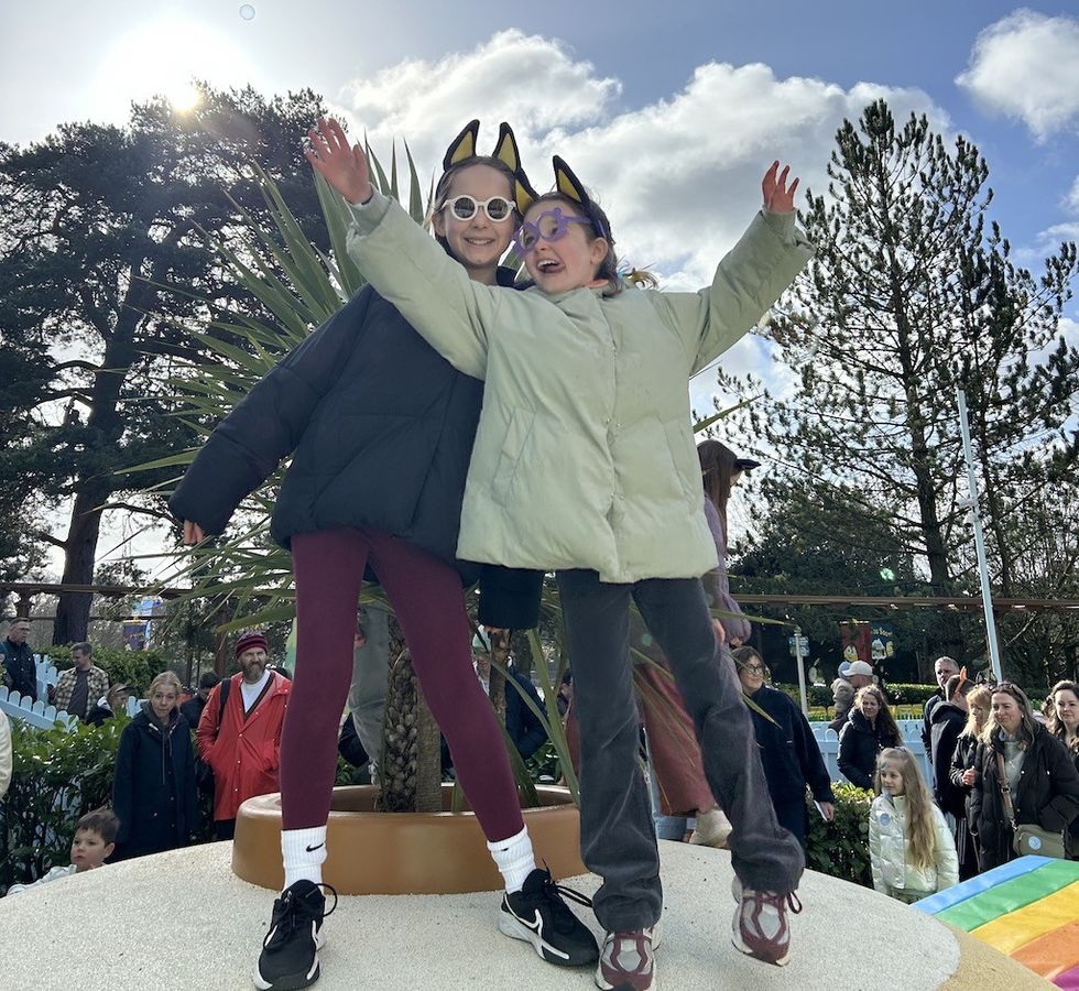 Two kids in jackets and fun sunglasses, arms raised, outdoor event with crowd and trees.