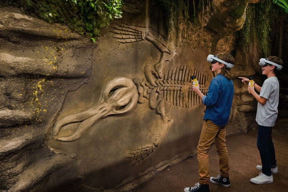 Two kids in VR headsets explore fossil wall in a museum exhibit.