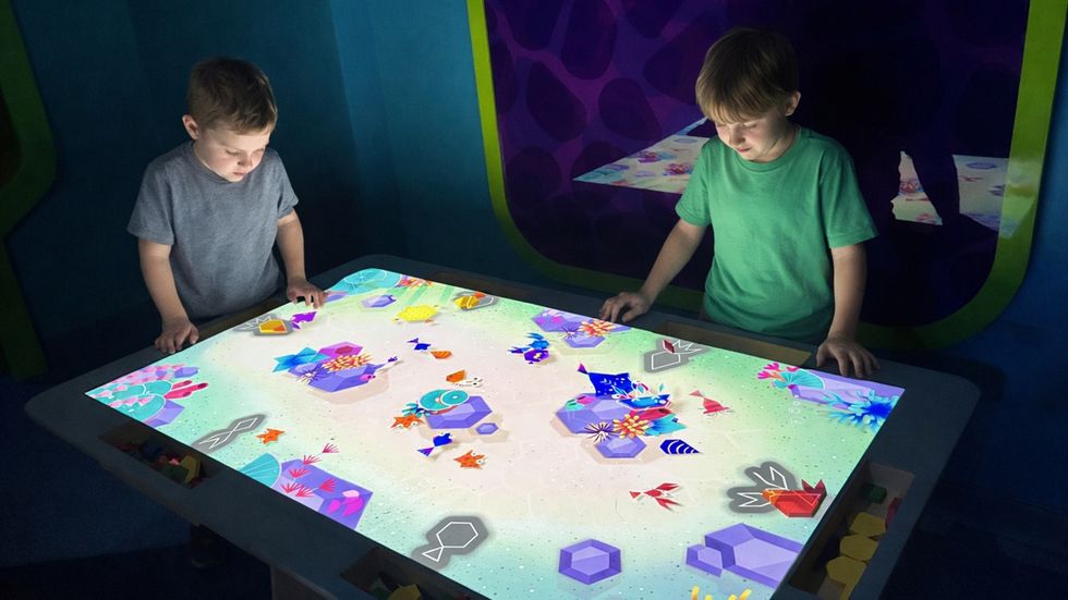 Two kids interact with a digital touchscreen table featuring colourful graphics and games form \u200bBreeze Creative