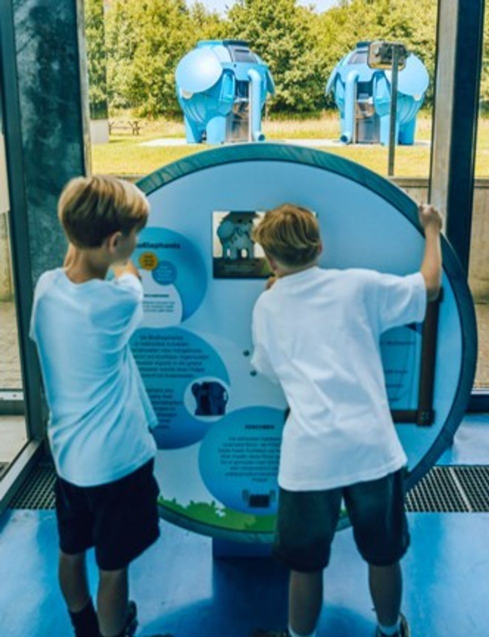 Two kids interact with an educational display, observing blue structures outside.