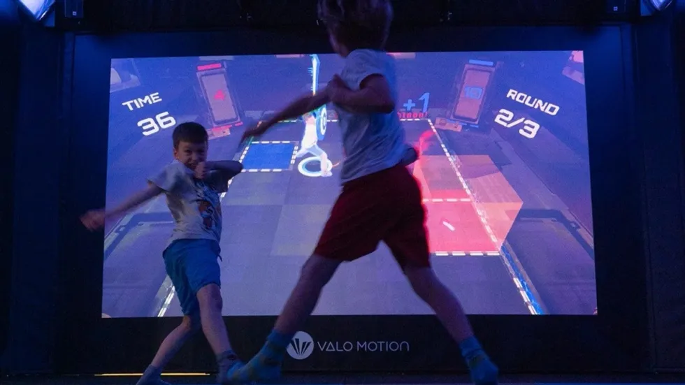 Two kids play an interactive game, moving energetically in front of a large digital screen.