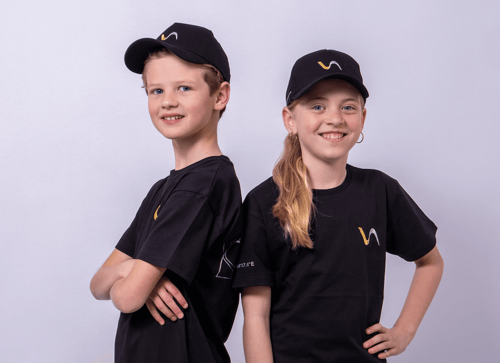 Two kids smiling, wearing matching black shirts and hats with Vekoma logos.