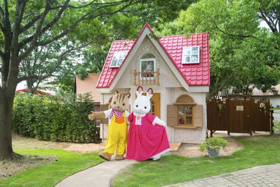 Two mascots in front of a small, charming house with a red roof in a garden setting.