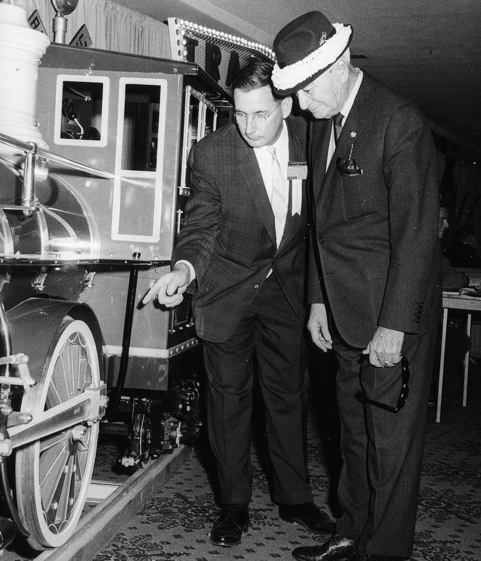 Two men examining a model train wheel in a showroom.