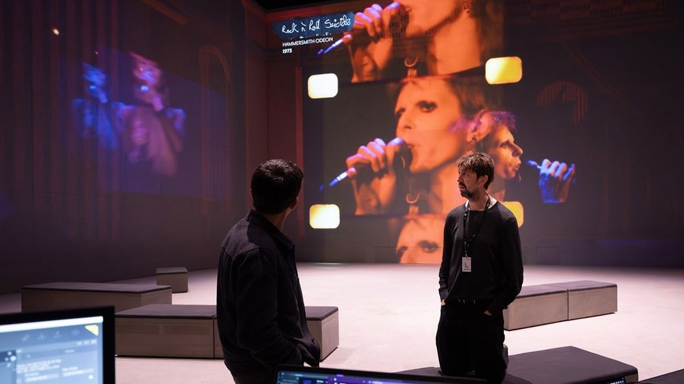 Two men in a room with large screens showing a singer performing.