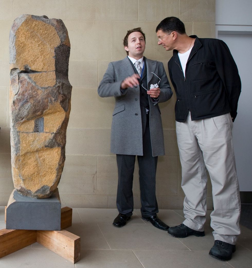 Two men in conversation beside a large stone sculpture indoors.
