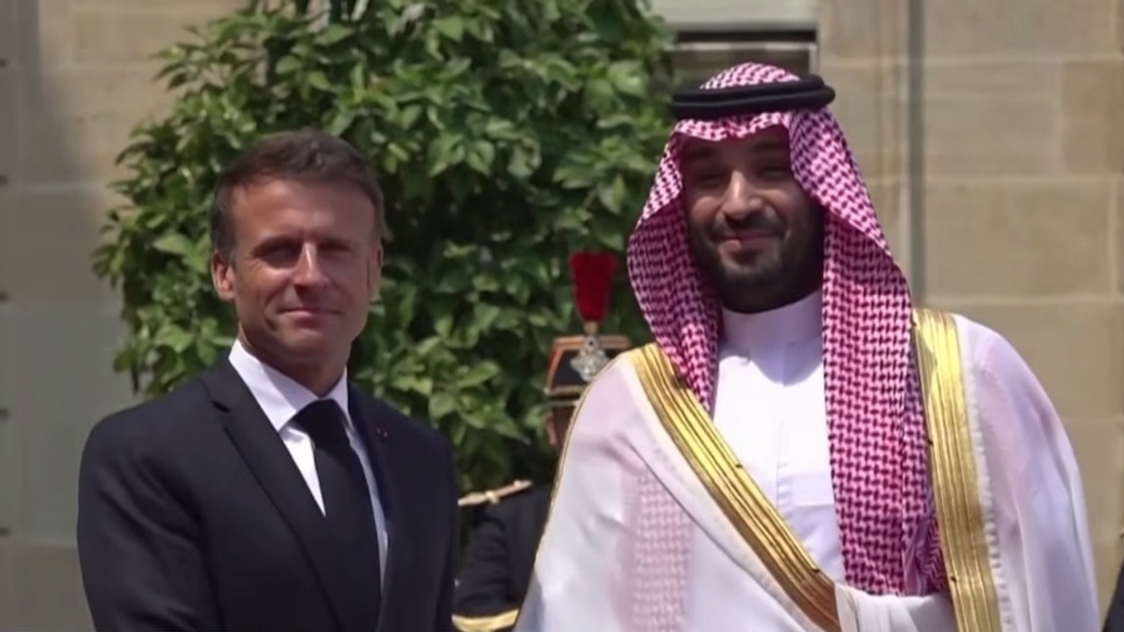 Two men in suits and robes, Saudi Arabia's Crown Prince Mohammed bin Salman and French President Emmanuel Macron, standing outdoors, smiling at a ceremonial event.