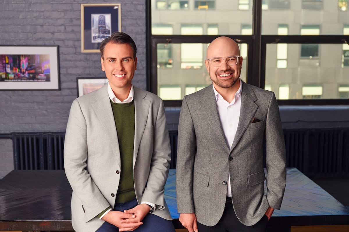 Two men in suits smiling in a modern office setting.