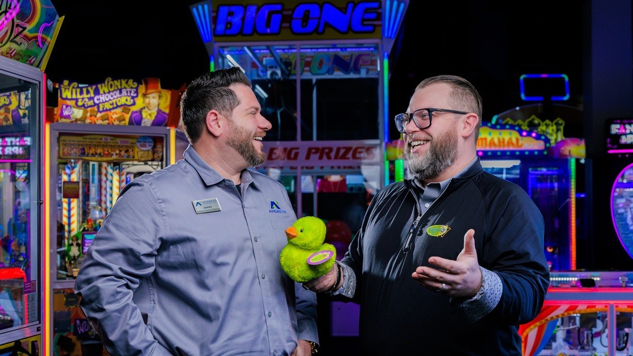 Two men laughing and holding a toy duck in a vibrant arcade setting.