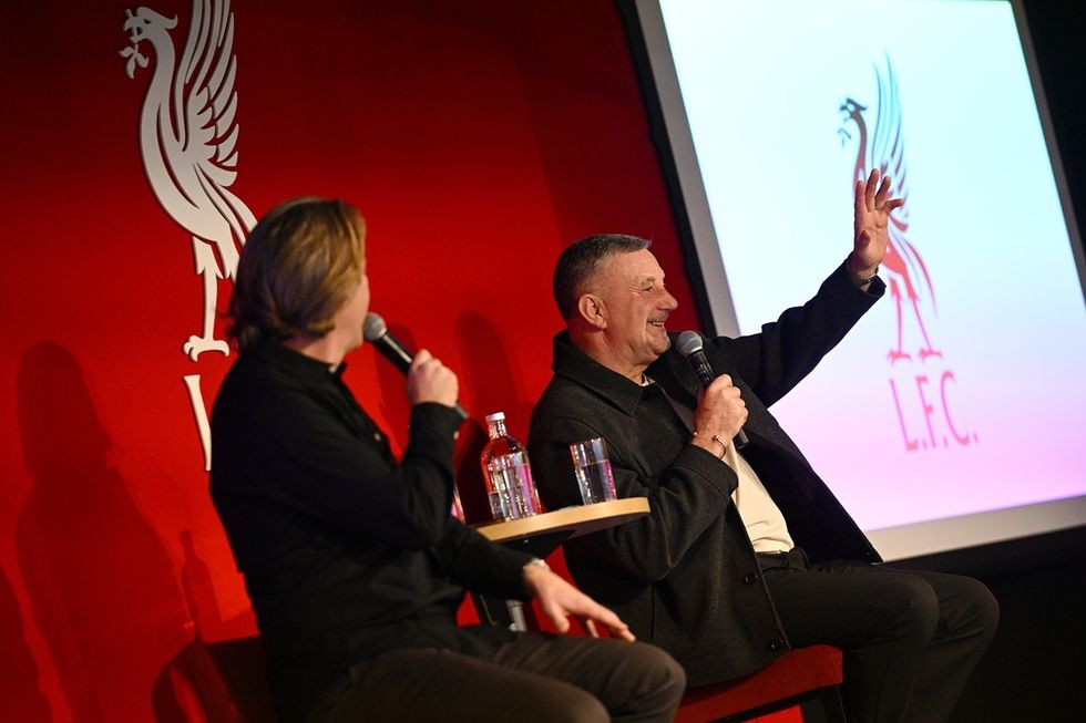Two men on stage, one waving, Liverpool F.C. logo in background.