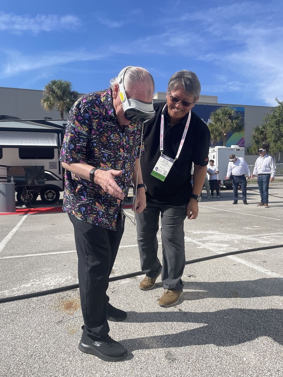 Two men outside, one wearing VR goggles and a colorful shirt, assisted by the other.