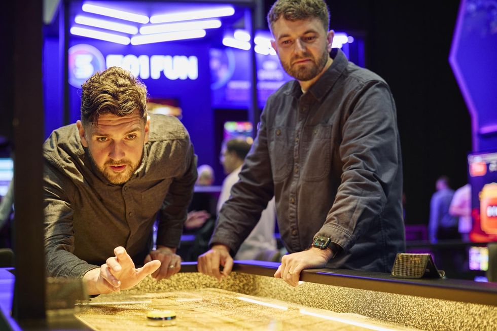 Two men playing shuffleboard in a neon-lit gaming area.