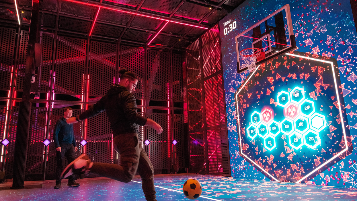 Two men playing Wall Ball on Glow Court, an interactive LED sports court aboard Norwegian Aqua. One player is mid-kick, aiming a yellow and black football towards a large digital screen displaying hexagonal targets, points, and a basketball hoop. The futuristic court is lit with vibrant neon red and blue lights, creating an immersive, tech-enhanced gaming experience on the ship’s top deck at sea.