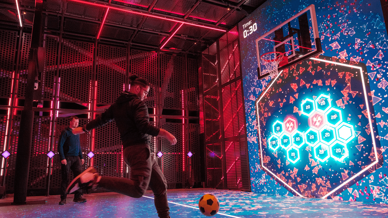 Two men playing Wall Ball on Glow Court, an interactive LED sports court aboard Norwegian Aqua. One player is mid-kick, aiming a yellow and black football towards a large digital screen displaying hexagonal targets, points, and a basketball hoop. The futuristic court is lit with vibrant neon red and blue lights, creating an immersive, tech-enhanced gaming experience on the ship’s top deck at sea.