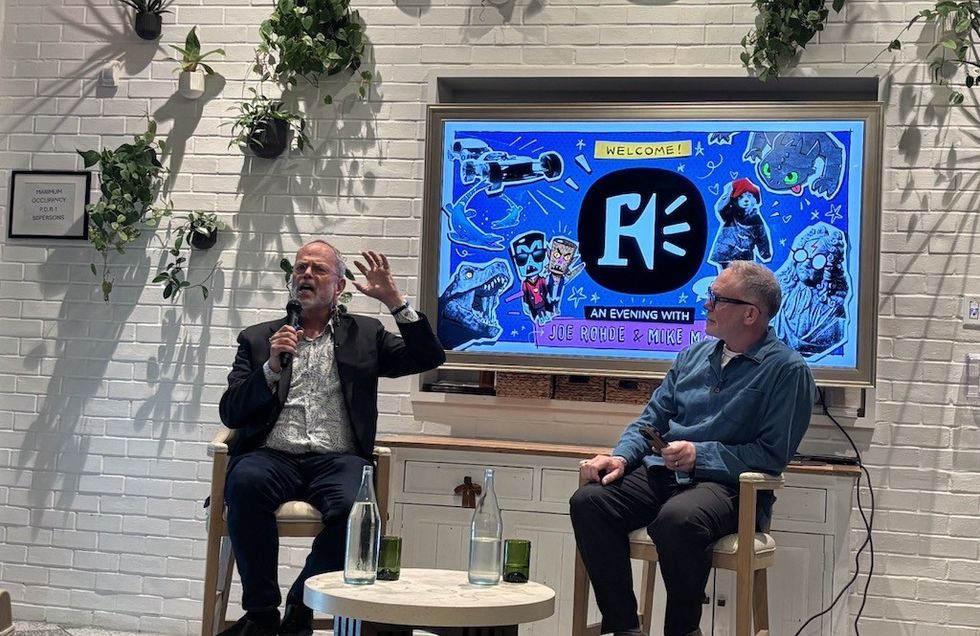 Two men seated in discussion, colorful screen backdrop, indoor plant decor.
