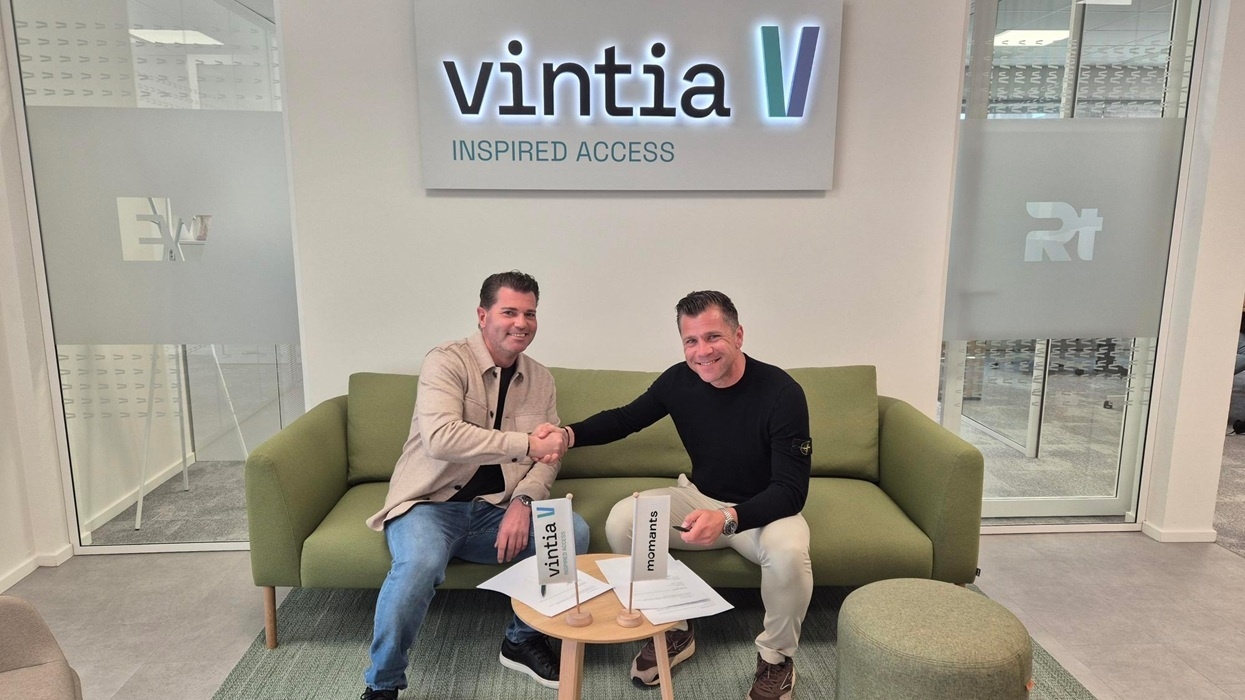 Two men shake hands in a modern office, sitting under a "vintia" sign, with flags for Vintia and Momants AI displayed on the table.